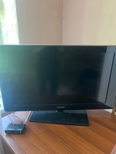 An indoor, eye-level, medium shot shows a Samsung television set with a digital set-top box connected to it, sitting on a wooden desk. The television is black with a thin bezel and a rectangular stand. The digital set-top box is small and black with a digital display showing "0347". The desk is made of polished wood. The background is a wall painted in a pale peach color, with a window on the left casting a bright light. The overall scene suggests a domestic setting. The image was taken in Berd, Armenia.