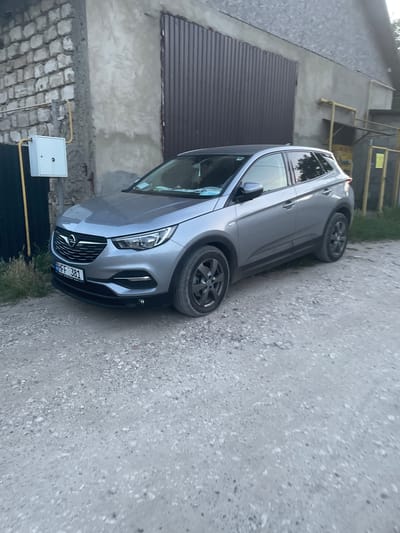 This media file captures a gray Opel Astra parked on a gravel surface. The car is angled, with its front facing towards the right of the frame. The vehicle appears to be in a somewhat utilitarian setting, possibly a rural or industrial area, with a building constructed of brick and concrete visible in the background. A brown corrugated metal door or panel is present on the building's exterior, and a white electrical box with yellow conduits is mounted on the wall.
The lighting suggests it is daytime, though the overall atmosphere is somewhat muted, implying it could be overcast or late afternoon. The car itself is the primary subject, with details like its license plate clearly visible. The license plate displays the text "HFF 381". The scene is static, with no people or apparent activity taking place. The car's wheels are dark gray alloy rims. The backdrop includes elements of urban or semi-urban infrastructure, including scaffolding-like structures and what appear to be utility lines. The ground is primarily composed of gravel and dirt.