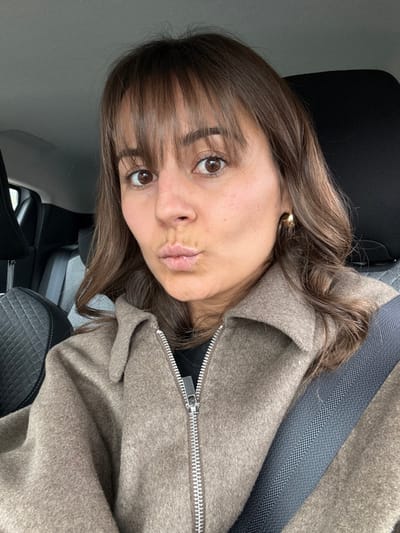 A female individual is depicted in a close-up portrait, occupying the majority of the frame, inside a vehicle in Mougins, France. She has brown hair with bangs and wavy shoulder-length strands, and brown eyes. Her lips are puckered in a "kissing" gesture, and she looks directly at the camera. She wears a light brown or taupe-colored collared jacket, partially open, with a prominent silver zipper. A dark garment is visible underneath the jacket. A gold-colored hoop earring is present on her visible right ear. A dark gray vehicle seatbelt diagonally crosses her chest and shoulder. The background reveals portions of a vehicle interior, including dark-colored car seats, one with a quilted pattern on the headrest.