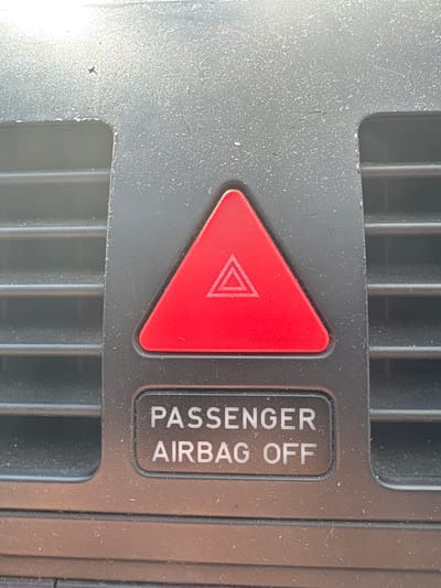The image shows a close-up of a car's dashboard. In the center, there is a red hazard light button, which is triangular with a white outline of a triangle inside. Below the hazard light button is a rectangular label with white text that reads "PASSENGER AIRBAG OFF". The dashboard is made of dark grey plastic and has horizontal vents on either side of the buttons. The surface of the dashboard appears dusty. The image was taken in Grębanin, Poland, likely inside a parked vehicle.