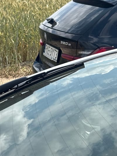 The media file shows the rear of a dark-colored BMW 320d parked on a grassy area, possibly a field, with dry, golden grass. The car's license plate is visible and reads "PKE 3YT3". A portion of the car's rear light is also visible. The car's rear window, which is reflective, shows a bright sky with clouds, suggesting it is daytime and likely clear or partly cloudy weather. A car windshield wiper is positioned in the lower right quadrant of the image, resting on the reflective rear window. The image appears to be taken outdoors, and the orientation suggests the photo was taken in a way that the car is not upright. The location context is Opatów, Poland. There are no people visible in the image.