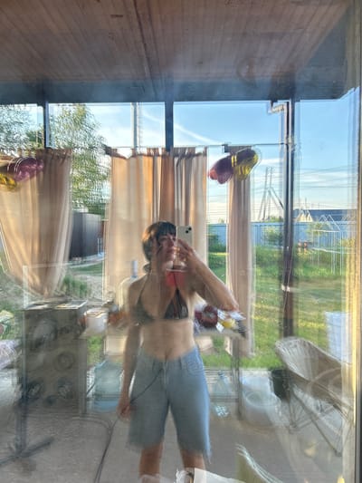 The image is a reflection in glass, appearing to be taken from inside a structure with a wooden ceiling and large glass panels, looking outwards. The reflection captures a young woman holding a smartphone, taking a selfie. She is wearing a dark patterned bikini top and light blue denim shorts. The background visible through the glass and in the reflection shows a suburban or rural environment, with green grass, trees, fences, and houses. There are also balloons, curtains, various objects on tables and shelves, and what appears to be a large water cooler bottle. The scene suggests a casual, outdoor setting possibly in a backyard or porch area, in the city of Pribrezhnyi, Russia, given the context provided. The lighting indicates it is daytime, possibly late afternoon with the sun casting shadows.