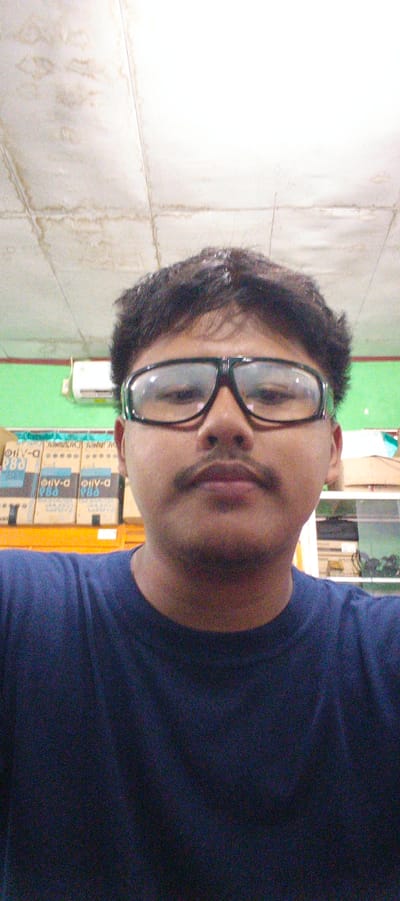 The image is a close-up, waist-up, selfie-style photograph of a young man wearing glasses and a dark blue t-shirt. He has dark, curly hair and a mustache. The background appears to be an indoor environment with a white ceiling with water stains, a green wall, and shelves with boxes. An air conditioning unit is visible on the green wall. The image quality is low, with visible noise and grain. The man's face is the primary focus, with the background out of focus. The location is in Sukabumi, Indonesia.