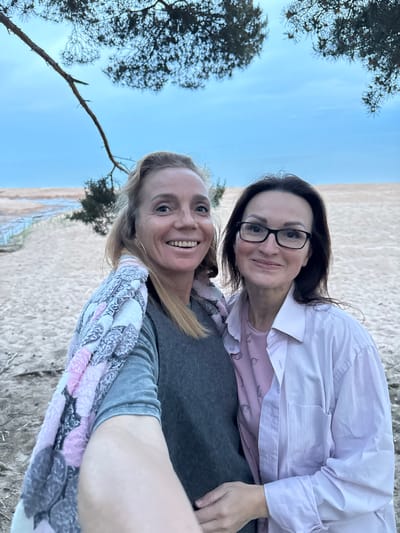 A selfie photograph captures two adult women on a sandy beach in Chernaya Lakhta, Russia. The woman on the left, with blonde hair and gold hoop earrings, is smiling broadly with teeth visible. She is wearing a grey long-sleeved top and has a multicolored (pink, grey, white, and black) patterned blanket draped over her left shoulder. Her right arm, extended forward, is visible at the bottom center, indicating she is holding the camera. The woman on the right, with dark brown hair and black-framed eyeglasses, is smiling gently with closed lips. She is dressed in a light pink collared button-up shirt worn over a darker pink patterned top. Her left hand is placed on the lower arm of the woman on the left. The background features light brown sand, with a narrow body of water and additional sandy banks visible on the left side of the frame. Sparse green vegetation, including a small bush and pine tree branches, is present in the mid-ground and at the top of the frame, respectively. The sky above is light blue with minimal cloud cover.