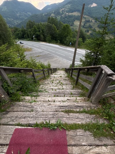 The image shows a view looking down a weathered wooden staircase that leads to a paved road winding through a mountainous, forested landscape. A dark-colored car is visible on the road, driving away from the viewer. The staircase is lined with a rustic wooden railing and is partially overgrown with green vegetation. In the foreground, a patch of dark red carpet is visible at the bottom of the stairs. The background features rolling green hills covered in dense trees, with some scattered houses visible on the slopes. The sky is overcast, with hints of sunlight breaking through the clouds. This scene is likely located in the vicinity of Mekića Kula, Montenegro, given the characteristic mountainous terrain and vegetation.