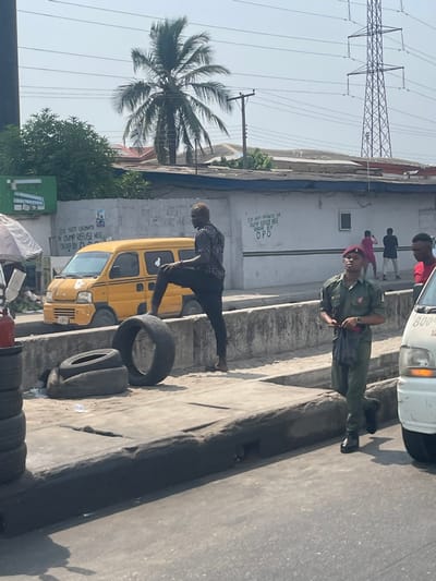 This image depicts an urban street scene in Awjaw, Nigeria, under clear daylight conditions. In the foreground, a stack of tires and a red fire extinguisher are visible on the left. A male subject with light-colored hair, wearing a dark patterned shirt and dark trousers, stands with his left foot elevated on a loose tire on a dusty, sandy surface adjacent to a concrete median strip. His right foot appears bare. Directly behind this individual, a yellow commercial mini-van is parked.
To the right, on the asphalt road, a male individual in a green military-style uniform, a red beret, and dark boots walks forward, looking slightly upward. He holds a dark object in his left hand. A portion of a white vehicle, displaying "800-" on its side, is visible on the far right of the road.
In the background, a light-colored building wall features black stenciled text: "DO NOT URINATE OR DUMP REFUSE HERE ORDER BY DPO." Several individuals are observed behind the concrete median and near this wall, one in a pink top. Above the buildings, a tall palm tree and dense green foliage are prominent. Overhead, numerous power lines stretch across a clear sky, with a large metal electricity transmission tower visible on the right.