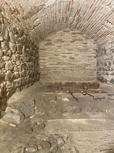 The image depicts the interior of a stone and brick vaulted chamber, likely a historical cellar or dungeon, located in Toledo, Spain. The walls are constructed of rough-hewn stones on the left and a mixture of brickwork and stones on the right and rear. The ceiling is a rounded brick arch, revealing exposed brick ribs. In the center of the chamber rests a weathered wooden structure, appearing to be a rack or primitive furniture, with metal straps attached to its front. The floor is uneven, covered in dirt, gravel, and larger stones. The environment appears aged and possibly part of an archaeological site or historical preservation area.