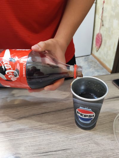 In Yalangach, Uzbekistan, a person wearing a red shirt is pouring Coca-Cola from a bottle into a Pepsi cup. The Coca-Cola bottle is red and white with the classic Coca-Cola logo and "CLASSIC" written on it. The Pepsi cup is black and white with the Pepsi logo. The scene takes place on a wooden table.
