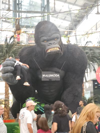 The image depicts a large, animatronic gorilla figure inside what appears to be a shopping mall in Perímetro Urbano Cúcuta, Colombia. The gorilla has exposed teeth and yellow eyes. A child is positioned in the gorilla's open hand. A "MALUMODA" logo is visible on the gorilla's chest. People are surrounding the gorilla. Artificial palm trees are in the background. The mall has a high ceiling. A man on a rope also appears to be a animatronic character.