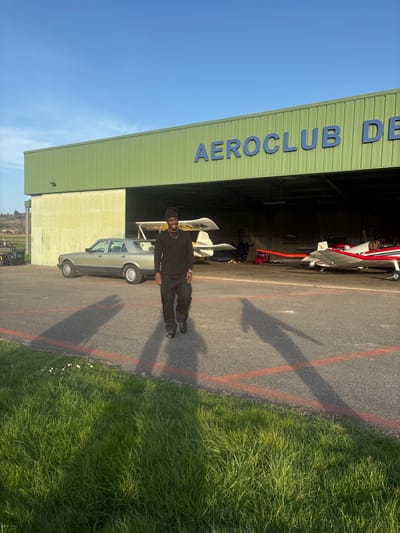 An outdoor scene at the Aéroclub de Chonas-l'Amballan, France, depicts a male subject walking towards the camera on an asphalt tarmac. The subject, a man with dark skin, is dressed in a dark beanie, dark long-sleeved shirt, dark pants, and dark shoes, exhibiting a slight smile. Long shadows, indicative of early morning or late afternoon sun, are cast from the subject and other objects across the asphalt, which is marked with two parallel red lines. The immediate foreground features green grass with small white flowers.
In the background, a large green and beige industrial-style hangar displays blue lettering spelling "AEROCLUB DE" on its upper facade. The hangar's large doors are open, revealing at least two single-engine aircraft inside: a white high-wing aircraft partially obscured on the left, and a white and red low-wing aircraft with registration F-PYP visible on its tail on the right. A light green/gold sedan is parked in front of the hangar, to the left of the man. Further left in the background, green fields, agricultural equipment, and rolling hills are visible under a clear blue sky.