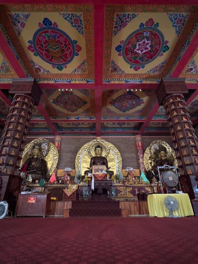 The image depicts the interior of a Buddhist temple in Việt Trì, Vietnam, likely the Trúc Lâm Đại Thiền Viện. The focal point is a central altar adorned with multiple Buddha statues, each with golden halos. The central Buddha statue is seated and appears to be holding a framed image. Flanking this central figure are two other Buddha statues, also seated and with elaborate halos.
The altar area is richly decorated with colorful textiles, offerings, and smaller statues. Behind the altar, the wall appears to be made of brick. The ceiling is a prominent feature, painted with intricate patterns and motifs in vibrant colors, including floral designs and circular medallions. Red wooden beams support the ceiling.
On either side of the altar are large, ornately carved wooden pillars that extend from the floor to the ceiling. These pillars are tiered and decorated with numerous small Buddha figurines. In front of the altar, the floor is covered with a deep red carpet with a subtle pattern.
To the left and right of the altar area, there are wooden structures that appear to serve as pedestals or display cases. On the left, a wooden cabinet with a sign that reads "HOM CÔNG ĐỨC" (meaning "Donation Home" or "Merit Home" in Vietnamese) is visible, with a fan in front of it. On the right, a table covered with a bright yellow cloth displays a large gong with a circular symbol in the center, and another fan is placed in front of it.
The overall impression is one of solemnity and grandeur, characteristic of a significant religious space. The lighting is somewhat subdued, contributing to the serene atmosphere.