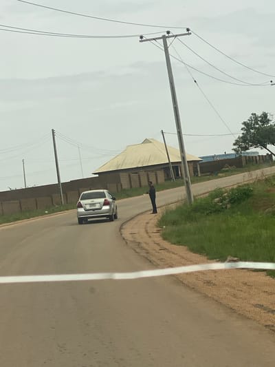 A silver hatchback car is driving on a paved road in a rural area. The car is facing away from the viewer and is in the center of the road. A person is standing on the side of the road next to the car. They are wearing dark clothing and appear to be facing the car.
The scene is an outdoor setting with a cloudy sky overhead, suggesting it could be morning or late afternoon. The road is lined with grass and some vegetation. There are utility poles with wires running overhead, indicating the presence of electricity infrastructure. In the background, there are buildings with light-colored roofs and brick walls. One building appears to be a large structure covered by a light-colored awning or tarp. The overall impression is a somewhat overcast day in a developing area, possibly a town or village. There is a white line painted on the road, marking the lane. No other specific location cues are immediately apparent beyond the general rural setting in DU, Nigeria. The visible text on the car's license plate is obscured. The person standing by the car has their back to the camera, making it impossible to discern any emotional expression.