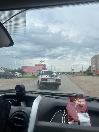 This media file captures a view from inside a vehicle, looking forward through the windshield. The scene depicts a street in Sorochinsk, Russia, with buildings and trees lining the road. Several cars are visible, including a white car in the foreground with a spider-like decal on its rear window and a license plate with Cyrillic characters. The sky is overcast with clouds, suggesting a possibly humid or rainy day, with a diffuse light indicating daytime. The vehicle interior is partially visible, showing a dashboard with a dashcam and a bag of snacks. The overall atmosphere appears to be that of a casual drive through a residential or semi-urban area. There is some text visible on the snack bag, which appears to be in Russian.