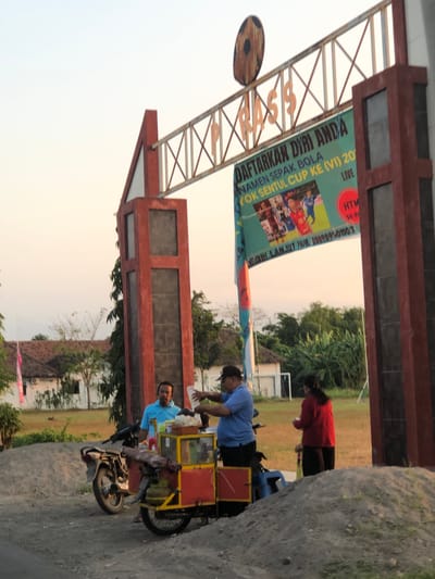 A mid-range shot captures a scene in the city of Sentul, Indonesia, with a clear sky in the background. In the foreground, a dirt pile sits on the right side of the image. To the left, a motorcycle is parked next to a food cart, which is also beside a man in a blue shirt. The food cart is red and yellow, with wheels and a gas tank. A woman in a red shirt stands behind the food cart. Further to the left, another man wears a blue shirt and a dark cap. A banner above the scene reads "DAFTARKAN DIRI ANDA" (Register Yourself) and mentions "YOK SENTUL CUP KE (VI) 20" with "LIVE" on the right. The banner also has a phone number and the words "LEBIH LANJUT HUB" (For more info, contact). Behind the people and cart are trees, buildings with tiled roofs, and a field. The overall scene appears to be outdoors, possibly near a sports field or event venue.