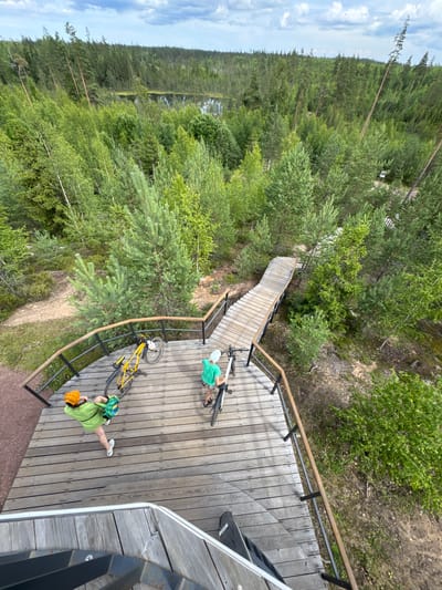 An overhead perspective shows a wooden viewing platform with two individuals and bicycles. On the left, a person wearing a green shirt and an orange helmet is leaning over a yellow bicycle. To the right, a person in a green t-shirt and a light-colored hat stands next to a bicycle. Both individuals are on the wooden platform, which features a railing with dark posts and wooden handrails. A winding wooden staircase extends from the platform into a dense forest area. The surrounding environment is a forest with various trees, including pines and birches, and a body of water is visible in the distance. The sky above is cloudy. The location is the city of Peski, Russia.