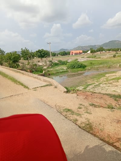 The image captures a rural landscape in Enagu, Nigeria, under a cloudy sky. In the foreground, a portion of a red object, likely a seat, occupies the lower left corner. A concrete road or bridge extends diagonally from the bottom left across the scene, with a thin strip of green grass along its edge. Beyond the road, a small body of water, a river or stream, meanders through a landscape of dry earth and sparse green vegetation. Trees are scattered across the middle ground, some appearing lush and green. In the background, a distinctive orange-colored building with multiple windows stands prominently. To its right, a collection of smaller buildings, appearing to be houses or structures, are visible. Further in the distance, rolling hills covered in greenery form the backdrop, with a few trees dotting their slopes. A utility pole with wires is visible in the middle distance, suggesting the presence of infrastructure. No people are clearly discernible in the image.