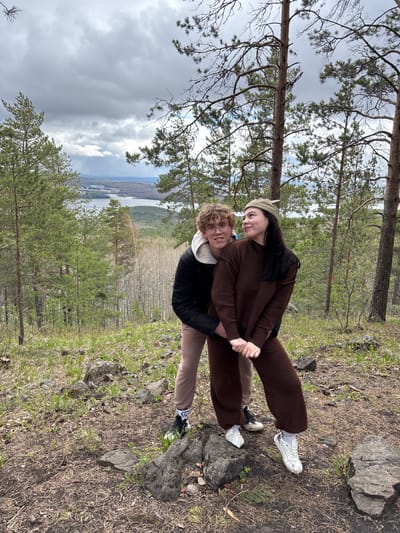 Two people are embracing outdoors. One person is wearing a brown sweater and pants, and a green cap. The other person is wearing a black jacket and light brown pants. They are standing on a hillside amongst trees and rocks. A body of water and city buildings are visible in the background. The location is in or near Kachkanar, Russia.