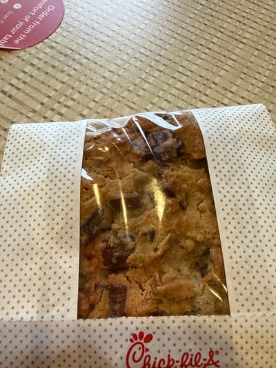The image shows a Chick-fil-A Chocolate Chunk Cookie in its packaging, sitting on a table. The cookie is rectangular and visible through a clear plastic window in the polka-dotted paper packaging. The Chick-fil-A logo is printed on the right side of the packaging. Part of a table service menu is visible in the bottom left.
While the exact location cannot be determined from this image, based on the presence of the Chick-fil-A cookie and table service menu, it can be inferred to be at a Chick-fil-A restaurant in Stone Mountain, United States of America.