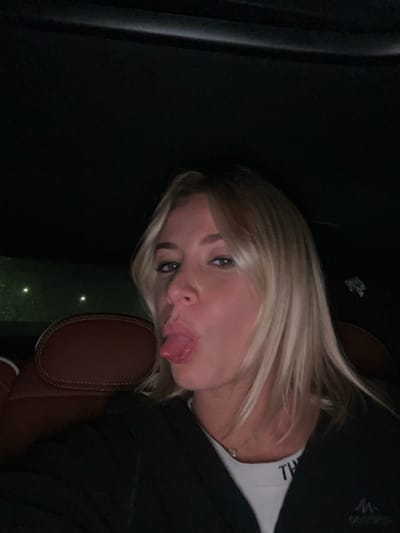 A blonde woman sticks her tongue out at the camera. She wears a white shirt and a dark jacket, and she has fair skin. The background is mostly black, with a brown car seat visible. The photo appears to have been taken in a car at night.