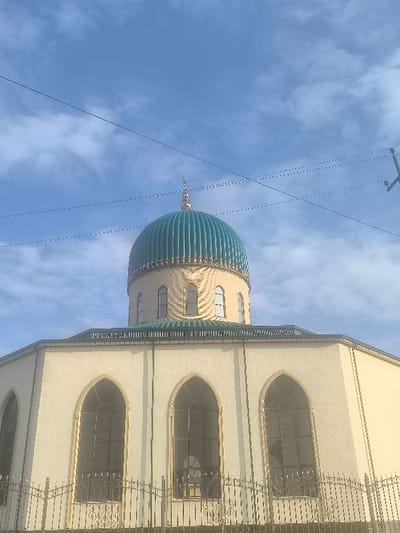 An upward-angled view captures a mosque in Dustlik, Uzbekistan, beneath a partly cloudy blue sky. The central feature is the mosque's large, ribbed, teal-green dome, crowned with a golden finial featuring a crescent moon. Below the dome, a cylindrical structure, light cream in color, exhibits multiple smaller, arched windows with light-colored frames. The main body of the building, also light cream, presents several larger, dark-framed arched windows. A decorative, wrought-iron fence runs along the lower visible section of the building's facade. Multiple dark utility wires traverse the upper portion of the sky, with a partial view of a utility pole structure visible on the far right. No individuals or specific actions are discernible.