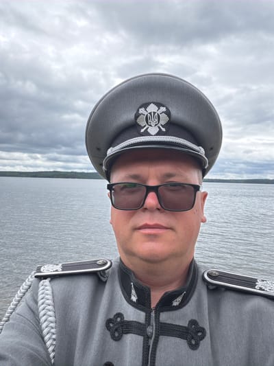 A man wearing a grey military-style uniform with a peaked cap stands in front of a body of water. The man has on dark-rimmed sunglasses. The uniform has silver braided cords on the shoulders and decorative black braiding on the front. The peaked cap is grey with a silver emblem. The background shows a cloudy sky and a distant tree-lined shore. The location is identified as Aneby, Sweden, specifically on the shore of Lake Ralången.