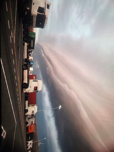 This image captures a dramatic sky over a truck parking lot, likely at Maasvlakte in Rotterdam, Netherlands. The sky is dominated by a striking shelf cloud, a sign of an approaching thunderstorm or squall line, casting an ominous and powerful atmosphere over the scene. The clouds are layered and textured, with a pinkish hue suggesting either sunrise or sunset light filtering through, or the light interacting with the storm's composition.

The foreground is filled with parked trucks, predominantly semi-trailer trucks with their cabs visible. Some trucks are hauling shipping containers. One prominent truck on the left has a green container with the text "EVERGREEN" clearly visible. Another truck further back displays "CAI" on its container, and one more has "Lloyd" on its orange container. These markings suggest the port or logistics nature of the location.

The parking lot is paved with asphalt and marked with white lines, indicating a structured area for vehicles. Lampposts are visible, suggesting the area is lit, and in the background, wind turbines can be seen against the stormy sky, a common feature of the Dutch landscape and a clue to the location. The overall mood is one of impending weather change, with the dramatic sky contrasting with the static presence of the trucks. There are no people visible in the frame, focusing the attention entirely on the landscape and the powerful natural phenomenon. The time of day is likely late afternoon or early evening, given the lighting conditions and the storm.