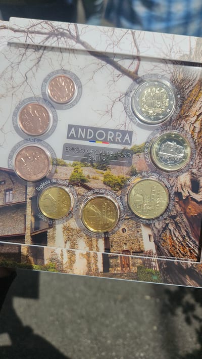 A rectangular commemorative blister pack of Andorran Euro coins from 2019 is displayed at a slight angle. The packaging features a photographic background of traditional stone buildings with multiple levels and visible window openings, set against a backdrop of trees, some with bare branches, and green conifers, indicating a mountainous or rural landscape. The words "ANDORRA," accompanied by a small Andorran flag (blue, yellow, red vertical stripes), are centrally printed, along with "EMISSIÓ OFICIAL DE L'EURO 2019."
Eight coins are individually encapsulated in transparent circular holders, revealing their obverse designs.
The three smallest copper-colored coins (1, 2, and 5 cents) depict an Isard (Pyrenean chamois) and are identified as "Acer recobert de coure" (copper-plated steel) with diameters of 16.25 mm, 18.75 mm, and 21.25 mm, respectively.
The three gold-colored coins (10, 20, and 50 cents) feature the Església de Santa Coloma (Church of Santa Coloma), a Romanesque church with a distinctive round bell tower, and are labeled "Or nòrdic" (Nordic gold) with diameters of 19.75 mm, 22.25 mm, and 24.25 mm.
The 1 Euro bimetallic coin, with a silver inner part and gold outer ring, depicts the Casa de la Vall (House of the Valley) and has a diameter of 23.25 mm.
The 2 Euro bimetallic coin, with a gold inner part and silver outer ring, displays the Escut d'Andorra (Coat of Arms of Andorra) and measures 25.75 mm in diameter. All coins are dated "2019."
The Església de Santa Coloma is located in the village of Santa Coloma, within the parish of Andorra la Vella, Andorra. The Casa de la Vall is a prominent historical building situated in the old town of Andorra la Vella, Andorra. The architectural style in the background image is consistent with structures found in the region of Andorra la Vella.
A partial view of a hand, with dark skin and a short sleeve, is visible in the bottom left corner, holding the coin set. The surface directly beneath the object is dark gray, textured, and out of focus, suggesting an outdoor ground surface such as asphalt or concrete.