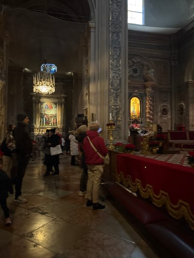 The image depicts the interior of the Ferrara Cathedral (Cattedrale di San Giorgio Martire) in Ferrara, Italy. The scene is a dimly lit, grand architectural space with individuals observing the surroundings.
Architectural features include high, vaulted ceilings, ornate fluted columns with intricate, gilded carvings, and arched doorways leading to various sections. Natural light enters from a high rectangular window on the right wall and a smaller, distinct blue stained-glass window high on the left. The floor consists of large, polished light-colored marble or stone tiles.
A prominent, multi-tiered crystal chandelier with numerous lit candles hangs in the mid-ground. Directly behind it, a large altarpiece features a painting depicting multiple figures within an ornate architectural frame. To the right, a side altar or niche contains a gilded, arched shrine housing an illuminated statue, likely a Madonna and Child. Golden candlesticks and arrangements of red poinsettias are visible on altars and other decorative surfaces. In the foreground, a long, dark red leather bench is present, fronted by a red velvet barrier with gold trim.
Approximately 10-15 individuals are dispersed throughout the visible area, standing and observing. Most are dressed in winter attire, including heavy coats and hats. A man in a dark jacket and beanie stands on the left, looking forward. A person in a bright red jacket and light-colored trousers stands in the mid-right foreground, facing away from the camera, holding a white smartphone or device at eye level, possibly photographing or recording. No direct interactions between individuals are discernible.