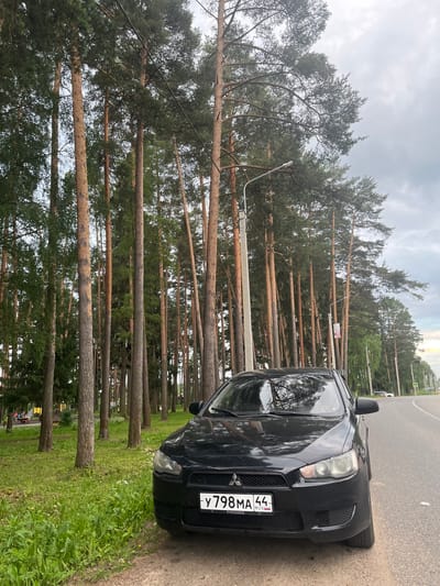 A black Mitsubishi Lancer is parked on the right side of a paved road. The car is angled towards the viewer, with its front end slightly turned to the right. The license plate reads "Y 798 MA 44" and "RUS". The car is positioned on a shoulder of the road, with a grassy area and a line of tall pine trees to its left. The trees are densely packed and extend high into the sky. The sky is overcast with a light gray hue. In the background, the road curves to the right, and a few other vehicles are visible in the distance. The overall environment suggests a rural or semi-urban setting.