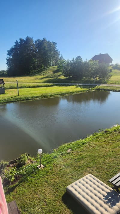 This is a horizontal, eye-level, full shot of a tranquil waterscape next to a well-maintained grassy area. The scene is bathed in the bright light of a sunny day, with a clear blue sky dominating the upper left portion of the frame. The sun's rays create a lens flare effect in the top left corner, indicating the time of day is likely late morning or early afternoon.

The main subjects are the still, calm water of a pond or narrow canal and the adjacent green grass. The water's surface is smooth, with subtle ripples reflecting the sky. The grass is neatly mowed, with distinct shadows cast by unseen trees, suggesting a well-kept outdoor space.

In the background, on the far side of the water, is a narrow paved path lined with trees that cast long shadows. Further back, a red-roofed building is visible, partially obscured by trees. On the right side of the frame, closer to the viewer, the grassy bank slopes down towards the water. A light-colored inflatable mattress is placed on the grass, suggesting a recreational or relaxation setting. A simple outdoor lamp with a white spherical head on a pole is also visible on the grass near the water's edge. The overall atmosphere is peaceful and idyllic, evoking a sense of calm summer weather. There is no visible text in the image. The location is identified as Lipuški, Latvia.