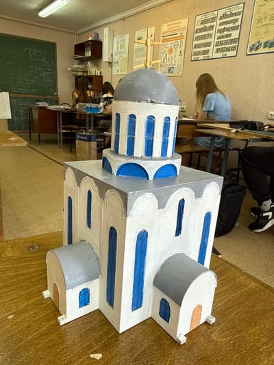 A detailed, close-up, eye-level, indoor shot features a handmade model of a church, likely a diorama or school project. The model is constructed from white and gray materials, with blue accents highlighting windows and architectural details. It sits on a wooden table in what appears to be a classroom or workshop setting. In the background, several students are visible, seated at desks and engaged in activities, suggesting a school environment. A blackboard with diagrams and writing is mounted on the wall behind them, along with various educational posters. The overall lighting suggests an indoor setting, possibly during daytime hours, given the fluorescent lights on the ceiling. The image does not provide any specific information or context that would allow for the determination of its location in Taganrog, Russia.