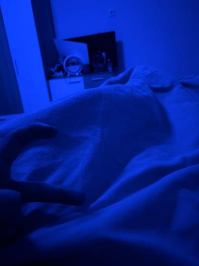 The image depicts an interior bedroom scene within the city of Nadarevo, Bulgaria, saturated with a dominant blue light source. In the lower-left foreground, a human hand with fingers curled is visible, partially obscured by light-colored bedding. The bedding, likely a duvet or sheet, occupies the majority of the foreground and midground, displaying various folds and creases under the blue illumination. The background, softly out of focus, features a tall, light-colored wardrobe on the left. Adjacent to it, a multi-drawer bedside table supports a circular object, possibly a clock or mirror, alongside other indistinct items. Above the bedside table, a dark, rectangular object, possibly a wall-mounted television, is visible. An electrical outlet is affixed to the wall in the upper-right portion of the background. The scene is static and appears to be under nocturnal or low-light conditions, with the blue light source strongly influencing the color perception.