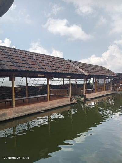 The image presents an outdoor, sheltered seating area constructed alongside a dark green water body, likely a pond or lake, in Palasari, Indonesia. A long structure with a multi-sectioned, brown-tiled roof extends horizontally across the frame, supported by numerous light-colored, vertical pillars. Beneath the roof, an elevated, hard-surfaced floor contains multiple dark benches and low tables, some featuring decorative carved panels. Two signs, labeled "Lara" and "Siapa," are affixed to overhead beams, indicating distinct seating sections. Clusters of small, light-colored decorative items are mounted on interior wall sections. The water surface exhibits ripples, reflecting the partially cloudy sky above, which features white cumulus formations against a light blue background. No individuals are present. The timestamp "2025/08/23 13:04" is visible in the lower-left corner.
