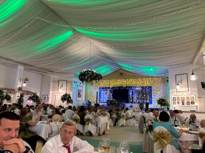 The image depicts an indoor event, likely a reception or party, taking place in the city of Vera, Spain. The ceiling is draped with white fabric, illuminated by green lights and strings of small white lights, creating a festive atmosphere. Several long tables are set for dining, covered with white tablecloths and adorned with floral arrangements and gold sashes on the chairs. People are seated at the tables, dressed in formal attire, suggesting a special occasion. Some individuals are engaged in conversation, while others are looking around or interacting with their surroundings. The background features a stage area with a large display of lights, possibly for entertainment. The overall scene conveys a sense of celebration and gathering.