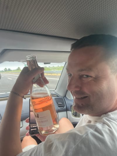 The image shows a close-up of a person's face as they look towards the camera. The person has short, dark hair, and is wearing a white shirt. They are holding a bottle of rosé wine, which is tilted as if to pour. The background appears to be the interior of a car, with a car's air vent visible, and a window showing a blurred landscape with trees and a road. The lighting suggests it is daytime, possibly overcast. A phone is also visible in the lower right corner of the image, held by the person. The label on the wine bottle is partially visible and reads "CO ROSÉ" and "2025". The overall tone is casual and candid.