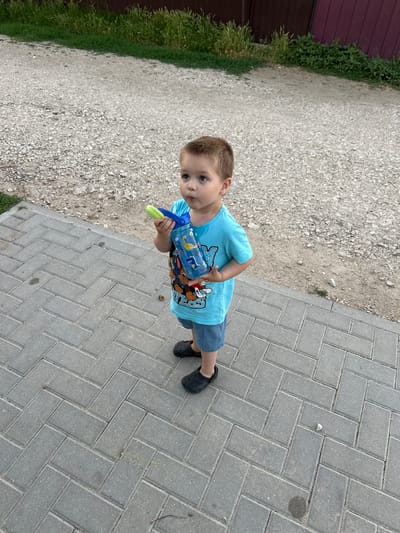 A young boy with brown hair and blue eyes is standing on a paved walkway, holding a blue and green water gun. He is wearing a blue t-shirt with a Paw Patrol graphic and blue shorts. The boy is looking towards the camera with a curious expression. To the left of the paved walkway is a gravel path, and further to the left is a dark red fence with green grass growing along its base. The paving stones are light grey and laid in a herringbone pattern. The weather appears to be overcast or it is late in the day, casting soft shadows. The setting seems to be an outdoor area, possibly a yard or a street in a village given the gravel and fence. There is text visible on the boy's t-shirt, which appears to be a Paw Patrol logo.