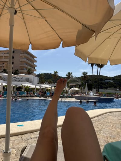The image depicts a resort setting with a swimming pool and a building. In the foreground, a portion of an umbrella and a person's legs are visible. The person's legs are in the lower right and center of the frame, with one leg extended and resting on the pool edge. The pool itself is a vibrant blue and is populated with several people swimming and relaxing. The background shows a lush green hillside with palm trees and a multi-story building, likely a hotel, with balconies. Umbrellas are scattered around the pool area, providing shade. The sky is clear and blue, suggesting a sunny day, likely late morning or early afternoon. The overall impression is one of a leisurely vacation day at a sunny resort. A small blue sign is visible on the edge of the pool, which reads "1.40 m".
