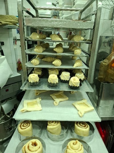 The image depicts a stainless steel rolling rack laden with various unbaked pastry items, situated within a commercial kitchen in Bnei Dror, Israel. The rack features multiple shelves, each holding different dough preparations on parchment paper. The top three visible shelves contain numerous unbaked croissants, arranged in rows. Below these, a shelf displays several unbaked pastries in fluted metal cups, some shaped like roses, alongside square-shaped pastries with raised edges and a central indentation. The lowest visible shelf holds multiple unbaked spiral pastries, resembling cinnamon rolls, each placed within an individual circular metal mold.

In the background on the left, a white tiled wall is visible, along with a stainless steel commercial stand mixer and a red fire extinguisher on the floor. A green quilted item rests on a shelf above the mixer. On the right, more white tiled wall and additional commercial kitchen equipment, including stainless steel containers and shelves, are partially visible. No people are present in the scene. The environment is consistent with a professional bakery or kitchen setting, showing prepared dough ready for baking.