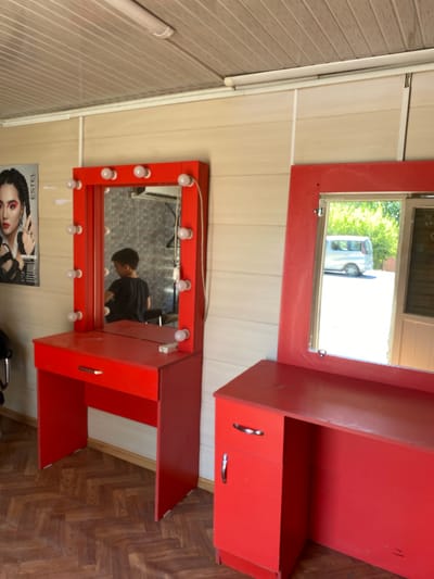 A boy with dark hair, wearing a black t-shirt, stands in front of a red vanity with a mirror. The mirror has lights around its frame. The vanity has a single drawer with a silver handle. The room has wood-paneled walls and a slatted ceiling with a fluorescent light fixture. To the left of the vanity is a poster of a woman with dramatic makeup. To the right is another red vanity with a mirror, this one showing a silver van parked in an outdoor area with green trees in the background. The floor is made of dark wood planks laid in a herringbone pattern. The location is identified as Konstantinivka, Kyrgyzstan.