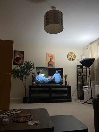 This image depicts a residential living room in Stevenage, United Kingdom. The room features cream-colored walls, a light ceiling with a central drum-shaped lamp featuring a geometric brown and black pattern, and a light-toned carpeted floor.
On the left, a light brown wooden door with a silver handle is visible, next to which hangs a rectangular abstract artwork with prominent circular shapes in orange, brown, and yellow tones. An artificial tree-like plant with green foliage in a black pot is positioned to the left of the central focus.
A large flat-screen television is centered on a black media console with glass-fronted compartments. The TV displays "Next Gen Chef, Episode 3: Wicked World Food," featuring two male individuals in white chef jackets; one on the left appears to be extending an arm towards the other, who has his hands open. On the console, a small globe is on the left, and a dark vase containing dried reeds is on the right.
Above the television, a rectangular artwork portrays a woman in an orange headwrap and dress, depicted in an African-inspired style. To the right, a large circular wall clock with a multi-colored, distressed face and black numbers is mounted. Further right, a black metal shelving unit contains multiple gold-colored decorative objects and artificial white orchids on its top shelf. A black softbox light on a tripod stands to the right of the shelving unit, next to a white portable air conditioning unit. A white radiator is visible on the far right, below light-colored curtains covering a window.
In the immediate foreground, two coffee tables are present. The upper table, dark brown with rounded corners, holds a white power adapter with a cable, a small wooden fan-shaped organizer, and a clear plastic bag containing small, reflective circular items. No individuals are physically present within the room.