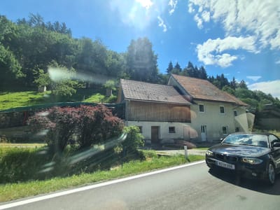 The image depicts a sunny day in Dürnstein in der Steiermark, Austria. A black convertible BMW is driving on a paved road in the foreground. Behind the car, a light-colored building with a tiled roof is visible, with a wooden annex attached to it. The building is situated on a grassy slope covered with trees and bushes. The sky is blue with scattered white clouds. The overall scene suggests a rural or suburban setting in Austria.