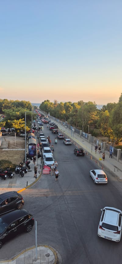 The image depicts a street scene in Paleo Faliro, Greece, during what appears to be late afternoon or early evening, given the warm lighting and clear sky. A considerable amount of traffic, composed primarily of cars and some motorcycles, is visible on a multi-lane road that curves gently. The road is flanked by a sidewalk on the left and a tree-lined area and a fence on the right.
On the left sidewalk, several people are standing or walking. There are also parked motorcycles and a white bus stop shelter. Some cars are parked along the left side of the road.
The road itself has traffic moving in both directions, with some congestion. Several pedestrians are visible on or near the sidewalk.
The right side of the road features a pedestrian walkway with a fence separating it from a densely wooded area. More people are seen walking on this side. In the distance, beyond the trees, the sea is visible, with a few boats or ships on the horizon.
The overall impression is that of a busy urban coastal area during a period of high activity.