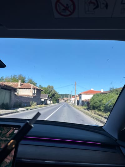 The image shows a view from inside a vehicle traveling on a paved road in the village of Veselinovo, Bulgaria. The road is flanked by traditional houses with red-tiled roofs and weathered facades. Green trees and vegetation line the sides of the road and surround the buildings, indicating a rural or semi-rural setting. The sky is clear and blue, suggesting a sunny day. The interior of the vehicle is visible in the foreground, with a dashboard and a digital display showing a map. A dark object, possibly a pen or stylus, is held in front of the screen. The overall scene depicts a journey through a Bulgarian village on a bright, clear day.