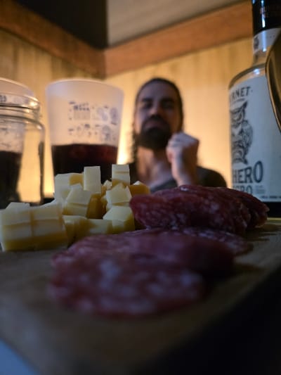 An indoor scene in Villa San Andrés, Argentina, displays a low-light setting with food and beverages. In the foreground, a wooden cutting board features an arrangement of approximately two dozen light-yellow cheese cubes on the left and multiple circular slices of reddish-pink cured meat on the right. In the mid-ground, from left to right, a clear glass jar containing a dark substance is partially visible, followed by a translucent white plastic cup holding a dark liquid and adorned with faint white illustrations. To the right of the cup, a dark glass bottle with a white label prominently displaying "FERNET" and "HERO" beneath an owl illustration is present. In the blurred background, a male figure with dark, medium-length hair and a beard is visible, wearing a dark shirt. His head is slightly tilted, and his right hand is raised, forming a loose fist near his chin or chest. The ambient lighting is dim, illuminating light-colored walls with a darker horizontal trim in the upper background.