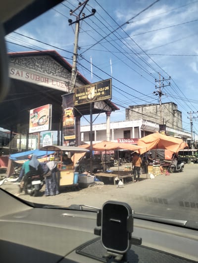 The image depicts a street scene in Keude Lueng Putu, Indonesia, likely a market area given the presence of stalls and vendors. In the foreground, a car's dashboard and windshield are visible, with a phone mount attached. The view through the windshield shows a busy street with several open-air market stalls covered by orange and blue tarps. People are seen interacting around the stalls, including a person wearing a blue headscarf and another in a blue garment, appearing to be patrons or vendors. A scooter is parked near a stall. In the background, there are buildings, including a structure with the sign "SAI SUBOH LUENG" and another with a yellow sign indicating "RAIDER KH..." and "KOMPI". Overhead, utility poles and a complex network of power lines dominate the sky, which is partially covered by clouds. The overall atmosphere suggests a vibrant daytime market in a commercial district.