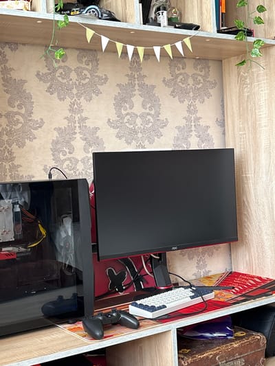 The image shows a desk setup within a room. The primary subjects are a computer monitor, a computer tower, a keyboard, and a gaming controller. The monitor is black with a red accent, and the AOC logo is visible on its bezel. The computer tower is also black with a transparent side panel, revealing internal components like fans and cables, including a "GEFORCE RTX" branded card. A white keyboard with blue and white keys is positioned in front of the monitor, with its cable connected. A black gaming controller rests on the right side of the desk.
The scene is set within a shelving unit with a light wood finish. The background features a beige wallpaper with a gray ornate pattern. There are decorative elements such as a string of small white and yellow pennant flags hanging on the left side of the shelving unit and a trailing artificial plant with green leaves. On the shelves, other objects are visible, including what appears to be a toy car, a bottle, and a vintage-looking suitcase on the right.
There is no clear indication of people, weather, or a specific time of day. The location context provided is Radkevshchina, Russia, but there are no explicit visual cues within the image to confirm this. The overall impression is of a tidy, functional, and personalized gaming or computer workstation.