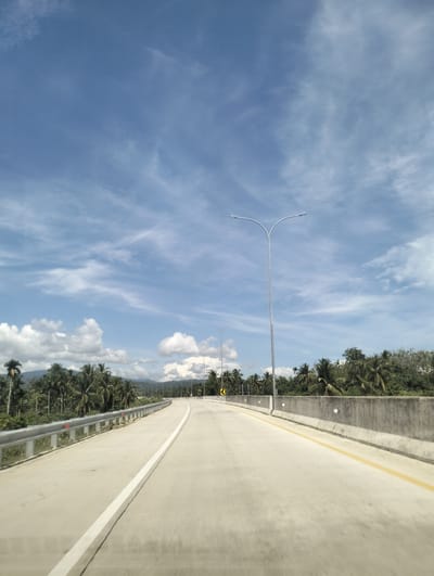 The image captures an elevated concrete highway winding through a lush tropical landscape. The sky above is a vibrant blue, dotted with scattered white clouds. On either side of the highway, palm trees and dense green foliage line the road, with mountains visible in the distance. The highway itself is marked with white and yellow lines, and several streetlights are mounted on tall poles along its edge. A metal guardrail runs along the left side of the road, and a concrete barrier is present on the right. The scene is devoid of any people or vehicles, suggesting a tranquil and unpopulated stretch of road. The overall impression is one of a modern infrastructure project integrated into a natural, tropical environment, likely in a location such as Mukee Gogo, Indonesia, characterized by its coastal or island geography and abundant vegetation.