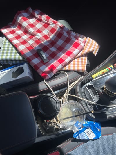 The image displays the interior of a vehicle, under daylight conditions, within the city of Wollongong, Australia. The scene focuses on the passenger side, revealing a collection of items on the seat and center console.
On the seat, a stack of folded checkered fabrics is present. The top fabric is predominantly red and white gingham. Beneath it, a green and white gingham pattern is visible, followed by a partially visible orange/tan and white gingham fabric. A dark grey, textured clip rests on the red gingham fabric. To the left, a white cardboard box, partially labeled "Anko Tape Dispenser," is visible.
The vehicle's center console features black upholstery with red stitching. On this console, a black disposable coffee cup with a black lid marked "CAUTION HOT" is situated next to a clear plastic cup containing a light-colored liquid and a dark straw. Multiple white cables are tangled between the cups and the gear selector area. A yellow Artline 70 marker pen with a black cap rests near the gear selector, which has a metallic ring at its base. A crumpled blue and white empty snack package, displaying a barcode and partial text including "Made in Australia," lies near the clear plastic cup. A segment of blue denim fabric is visible in the lower right portion of the frame, suggesting the presence or recent occupation of an adjacent seat. No individuals are directly visible.
