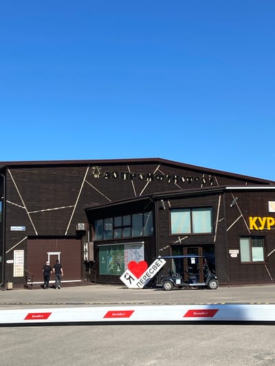 The image captures a modern, two-story building with dark brown horizontal siding and light-colored geometric accents, set against a clear blue sky on a sunny day. Large yellow Cyrillic letters, partially visible on the upper facade, spell "КУР" (likely part of "КУРОРТ" meaning "resort").

In the foreground, a white barrier arm, clearly branded "DoorHan" in red text, extends across a paved entrance. Two people, dressed in dark attire, are visible standing near an entrance to the building, partially obscured by the barrier. A small, dark-colored utility vehicle, resembling a golf cart, is parked near this entrance.

A prominent white sign, featuring a large red heart and the Cyrillic word "ПЕРЕСВЕТ" (Peresvet) followed by the letter "К", is positioned near the utility vehicle. This sign directly references the location context of Peresvet, Russia. Other smaller signs on the building include "ШВЕЙЦАРСКАЯ" (Swiss) and mentions of "SKI TRAS...", suggesting a recreational or resort complex. Security cameras are mounted on the building, and an air conditioning unit is also visible. The scene appears to be an access point to a facility during daytime.