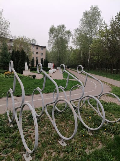 The image shows a public space in Uspenskoye, Russia. In the foreground, there are four metal bicycle racks shaped like bicycles, painted in light gray. The racks are positioned on a patch of grass and a paved surface with weeds and yellow flowers growing around them.
Behind the bike racks, there is a walking and bike path with a painted white line, indicating lanes. Further back, there is a lawn with sculptures and trees. A building with a light-colored facade and multiple windows is visible behind the trees. The sky is overcast, suggesting a cloudy day. A person is visible walking in the distance on the path.